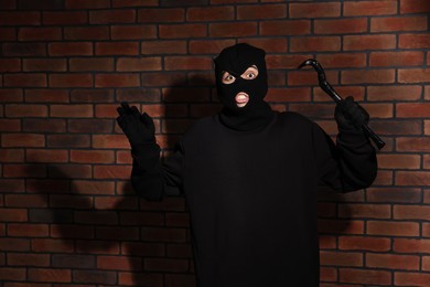 Emotional thief in balaclava raising hands with crowbar against red brick wall