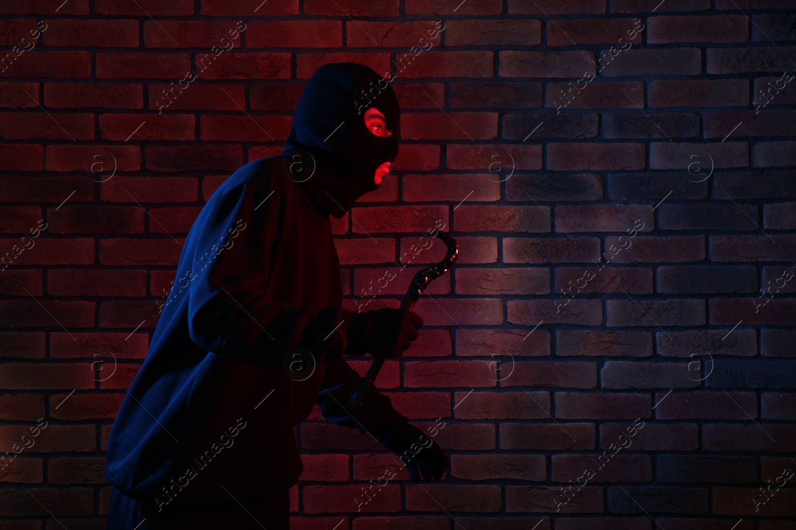 Photo of Thief in balaclava with crowbar in color lights against red brick wall. Space for text