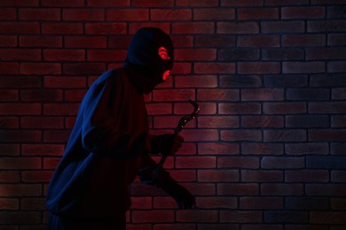 Photo of Thief in balaclava with crowbar in color lights against red brick wall. Space for text