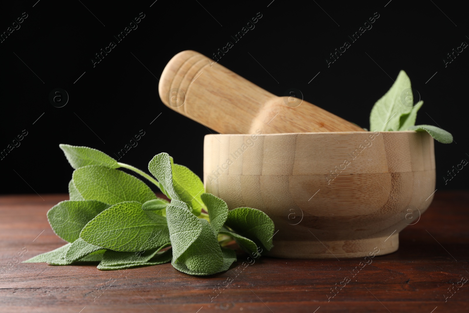 Photo of Green sage leaves, mortar and pestle on wooden table against black background, closeup