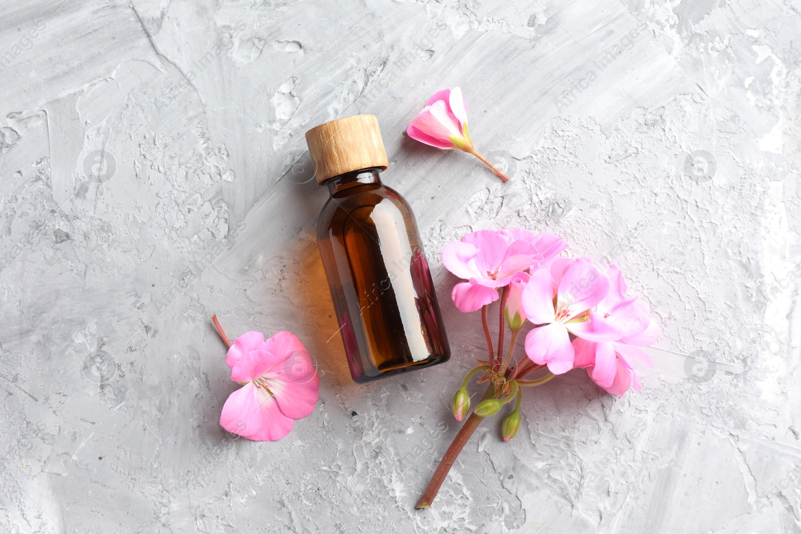 Photo of Bottle of geranium essential oil and beautiful flowers on light grey textured table, flat lay