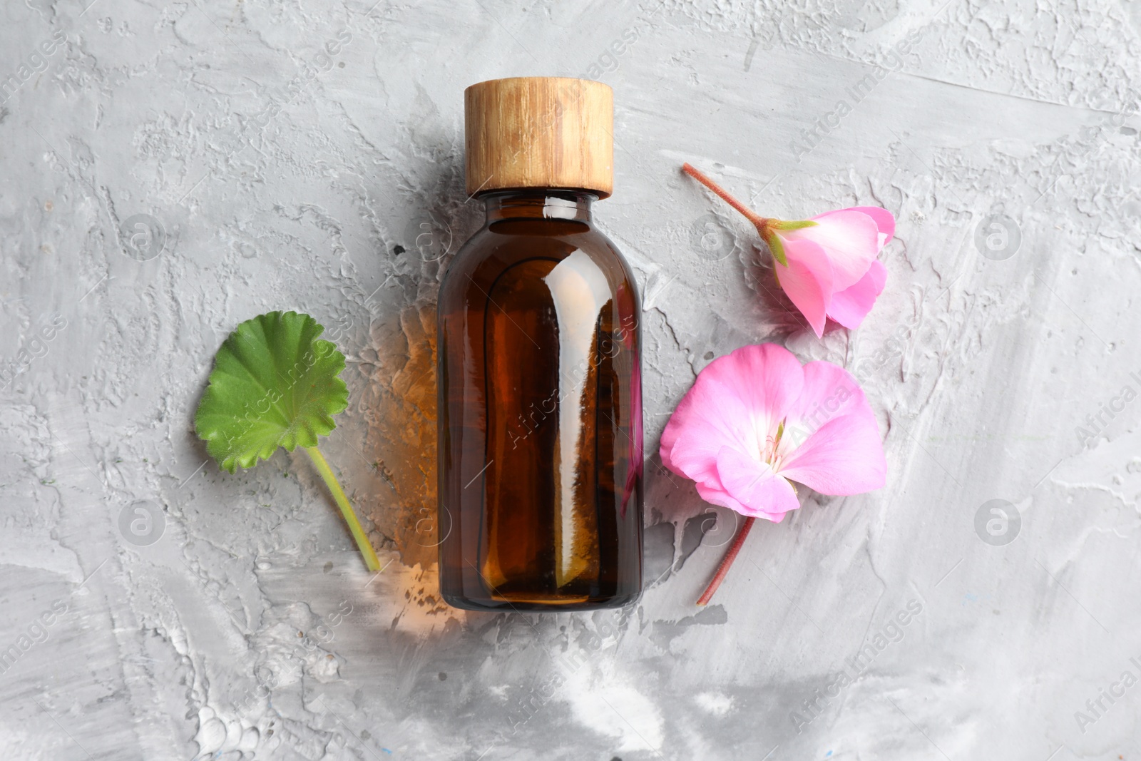 Photo of Bottle of geranium essential oil and beautiful flowers on light grey textured table, flat lay