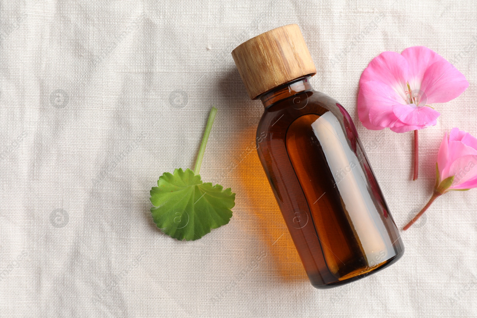 Photo of Bottle of geranium essential oil and beautiful flowers on white fabric, flat lay. Space for text