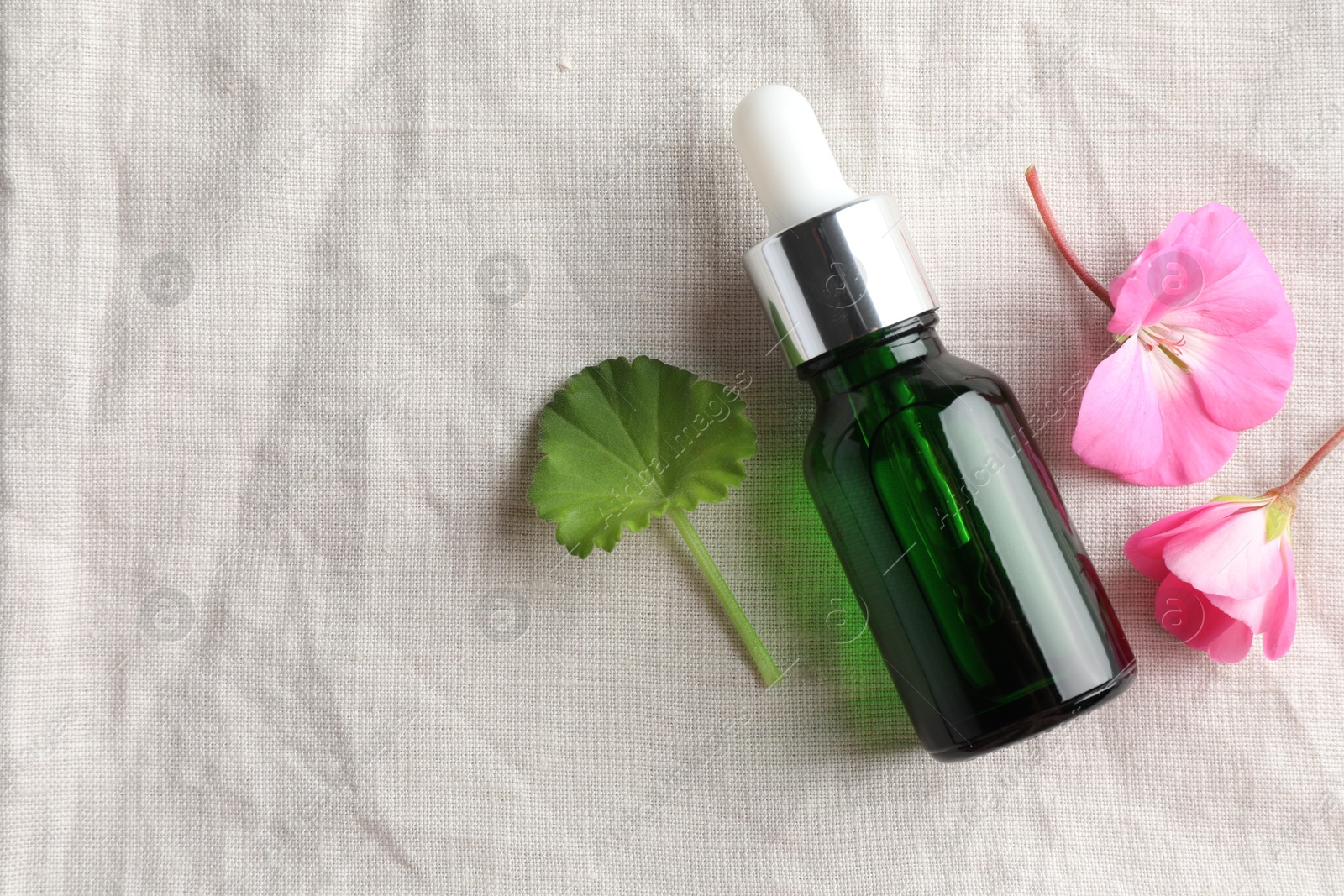 Photo of Bottle of geranium essential oil and beautiful flowers on white fabric, flat lay. Space for text