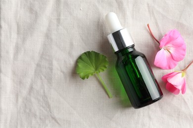 Photo of Bottle of geranium essential oil and beautiful flowers on white fabric, flat lay. Space for text