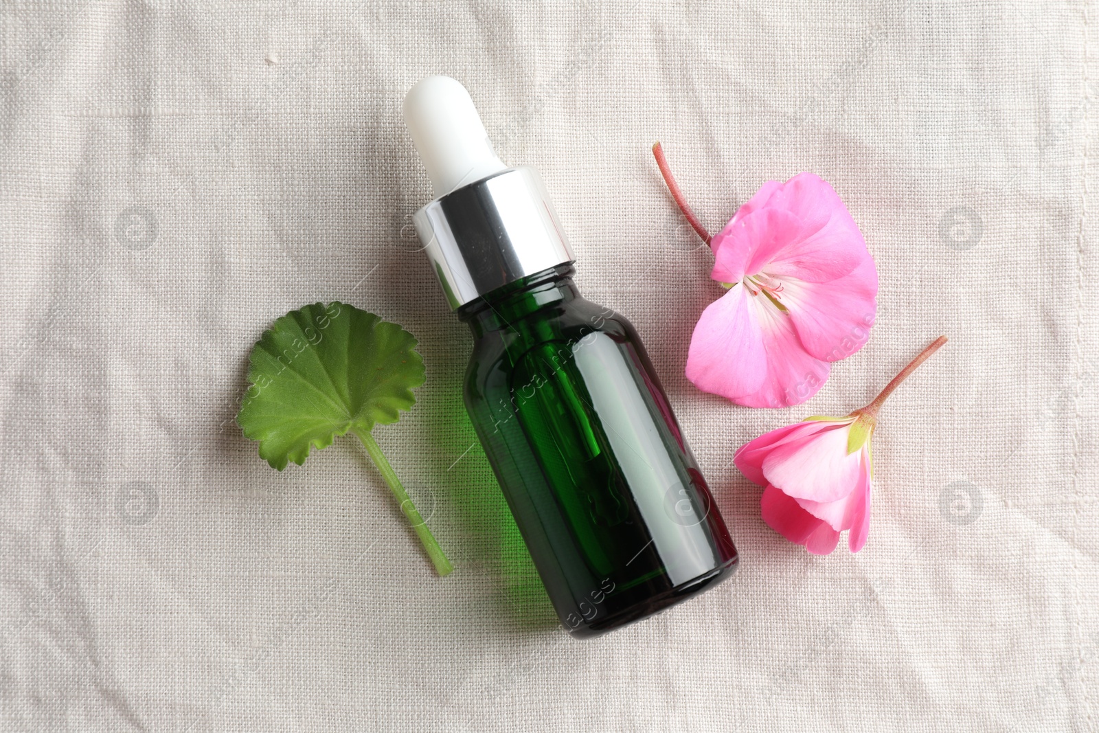 Photo of Bottle of geranium essential oil and beautiful flowers on white fabric, flat lay