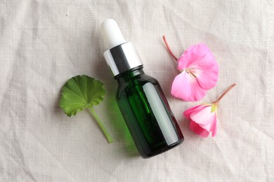 Photo of Bottle of geranium essential oil and beautiful flowers on white fabric, flat lay