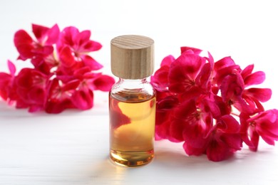 Photo of Bottle of geranium essential oil and beautiful flowers on white wooden table, closeup