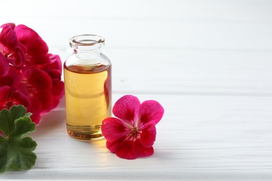 Photo of Bottle of geranium essential oil and beautiful flowers on white wooden table. Space for text