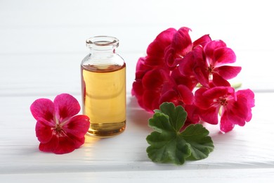 Photo of Bottle of geranium essential oil and beautiful flowers on white wooden table, closeup