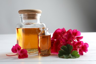 Photo of Bottles of geranium essential oil and beautiful flowers on white wooden table, closeup