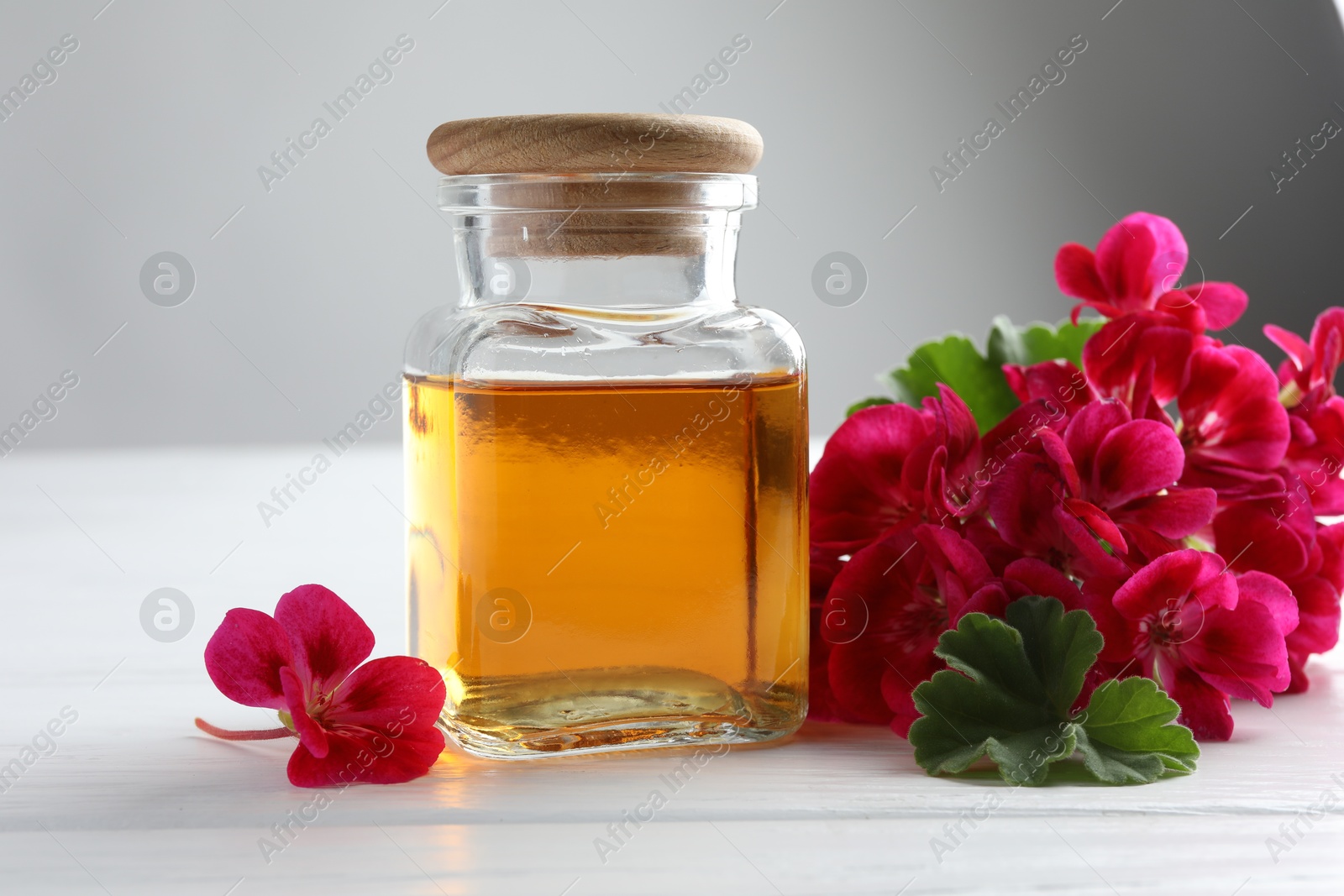 Photo of Bottle of geranium essential oil and beautiful flowers on white wooden table, closeup