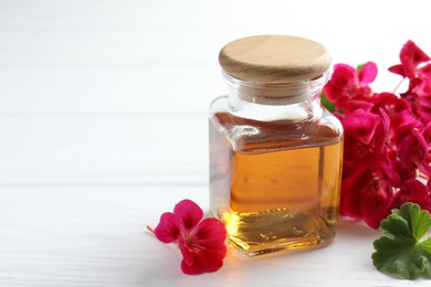 Photo of Bottle of geranium essential oil and beautiful flowers on white wooden table, closeup. Space for text