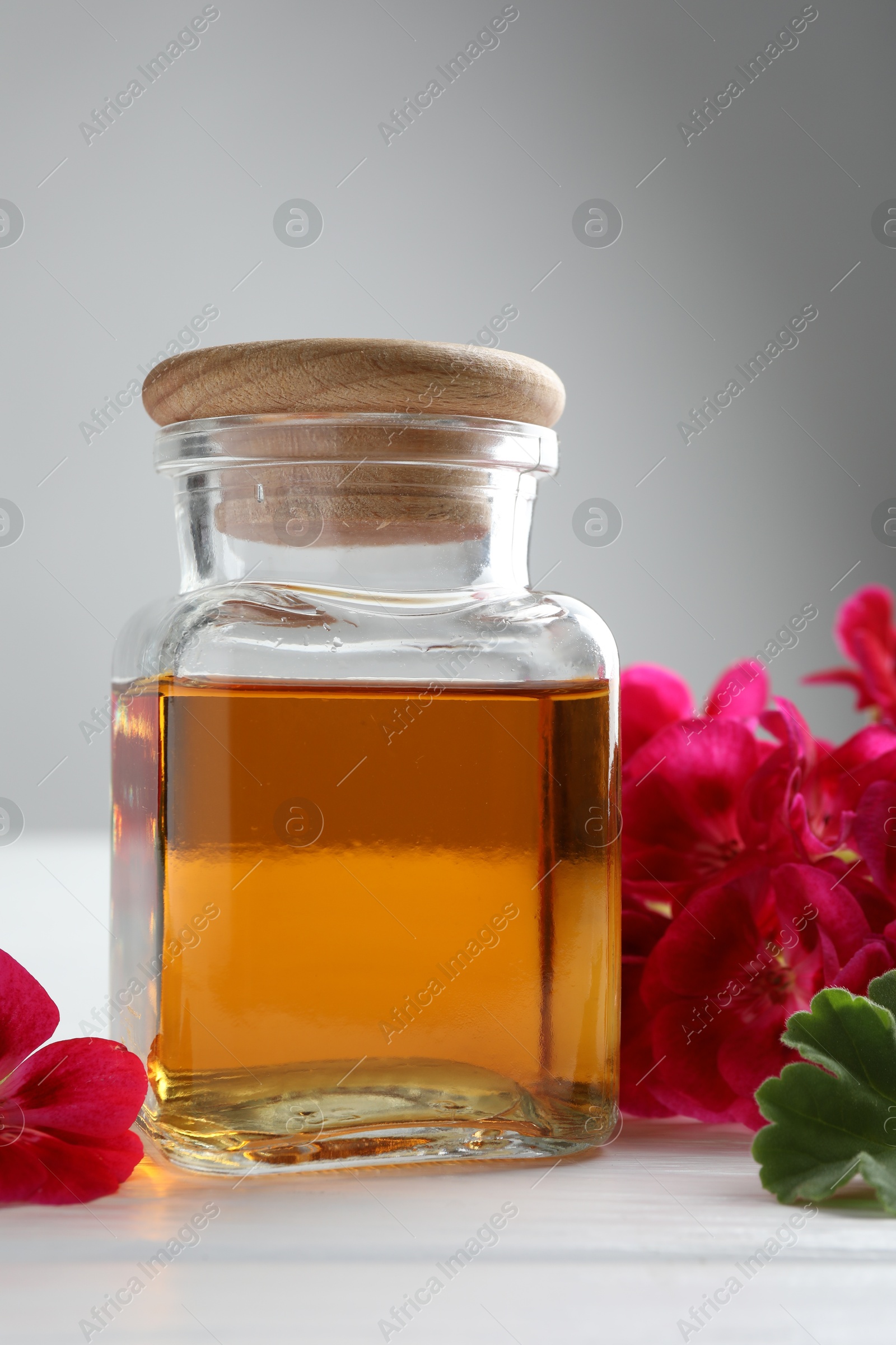 Photo of Bottle of geranium essential oil and beautiful flowers on white wooden table, closeup