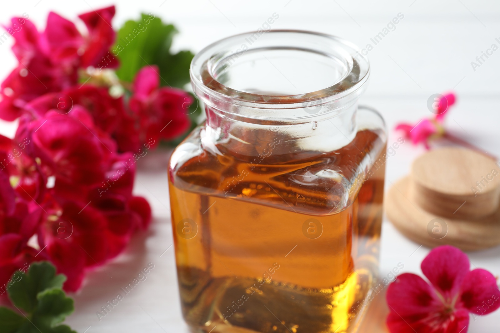 Photo of Bottle of geranium essential oil and beautiful flowers on white table, closeup