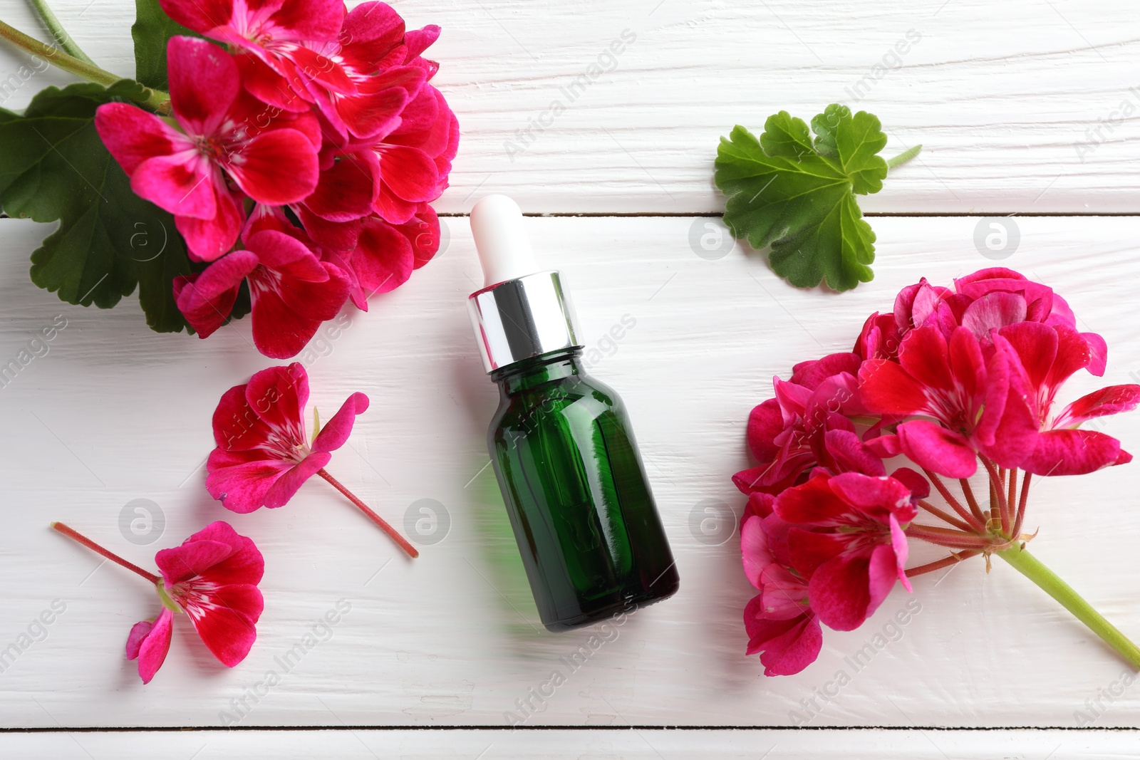 Photo of Bottle of geranium essential oil and beautiful flowers on white wooden table, flat lay