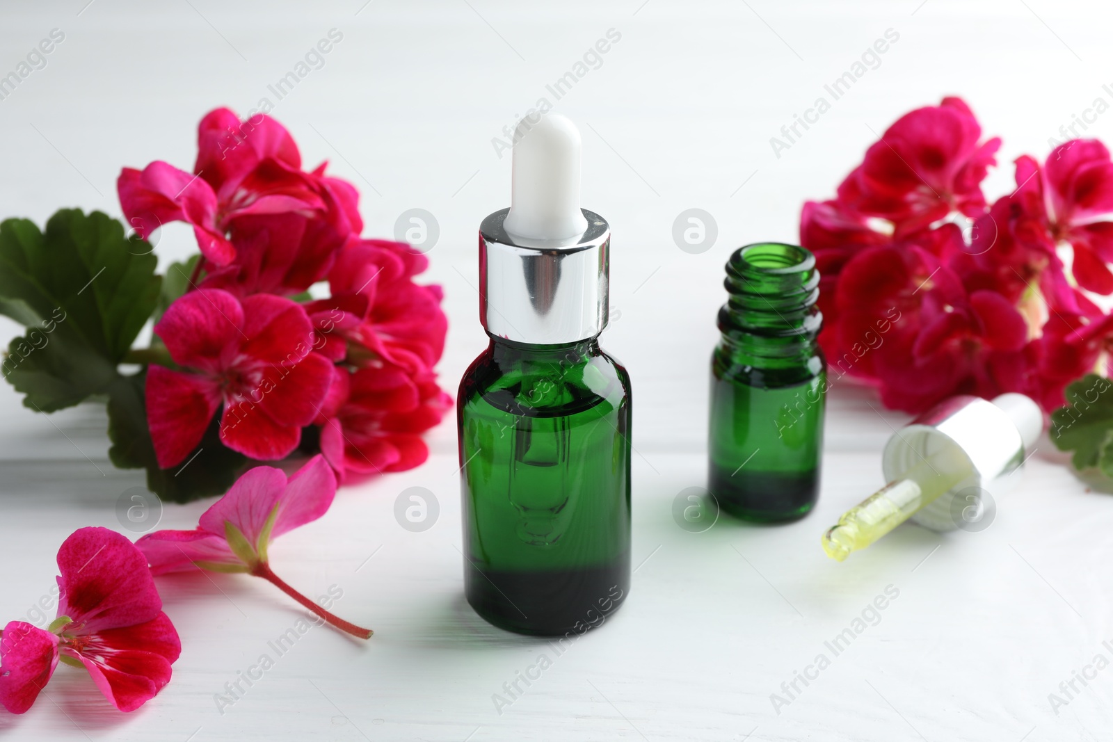 Photo of Geranium essential oil, pipette and beautiful flowers on white wooden table, closeup