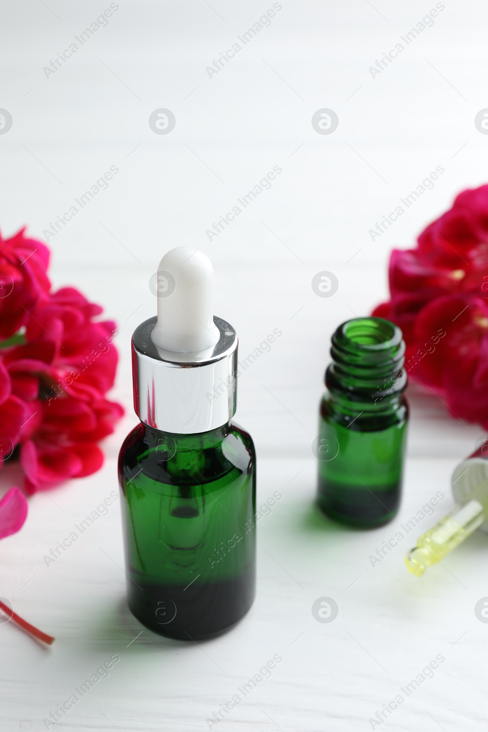 Photo of Geranium essential oil, pipette and beautiful flowers on white wooden table, closeup