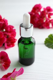 Photo of Bottle of geranium essential oil and beautiful flowers on white wooden table, closeup