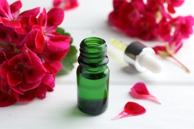 Photo of Bottle of geranium essential oil and beautiful flowers on white wooden table, closeup