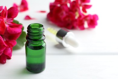 Photo of Bottle of geranium essential oil and beautiful flowers on white wooden table, closeup. Space for text