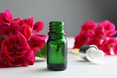 Photo of Bottle of geranium essential oil and beautiful flowers on white wooden table, closeup