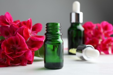 Photo of Bottles of geranium essential oil, pipette and beautiful flowers on white wooden table, closeup