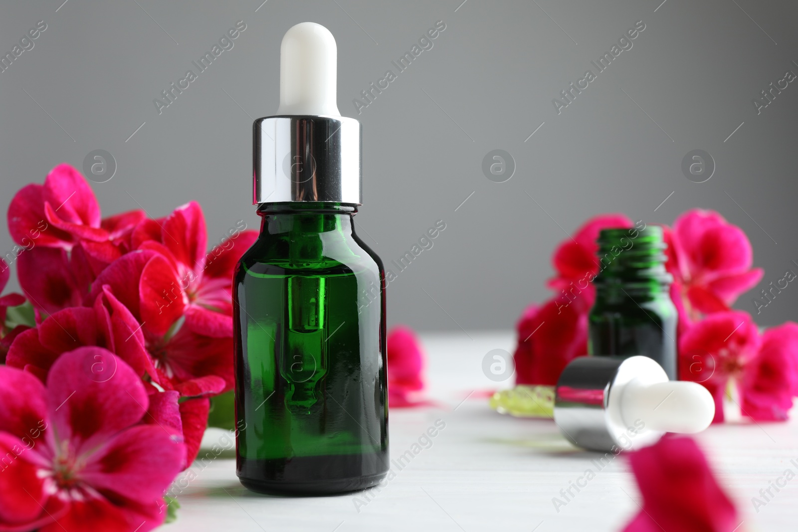 Photo of Bottles of geranium essential oil, pipette and beautiful flowers on white wooden table, closeup