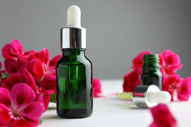 Photo of Bottles of geranium essential oil, pipette and beautiful flowers on white wooden table, closeup