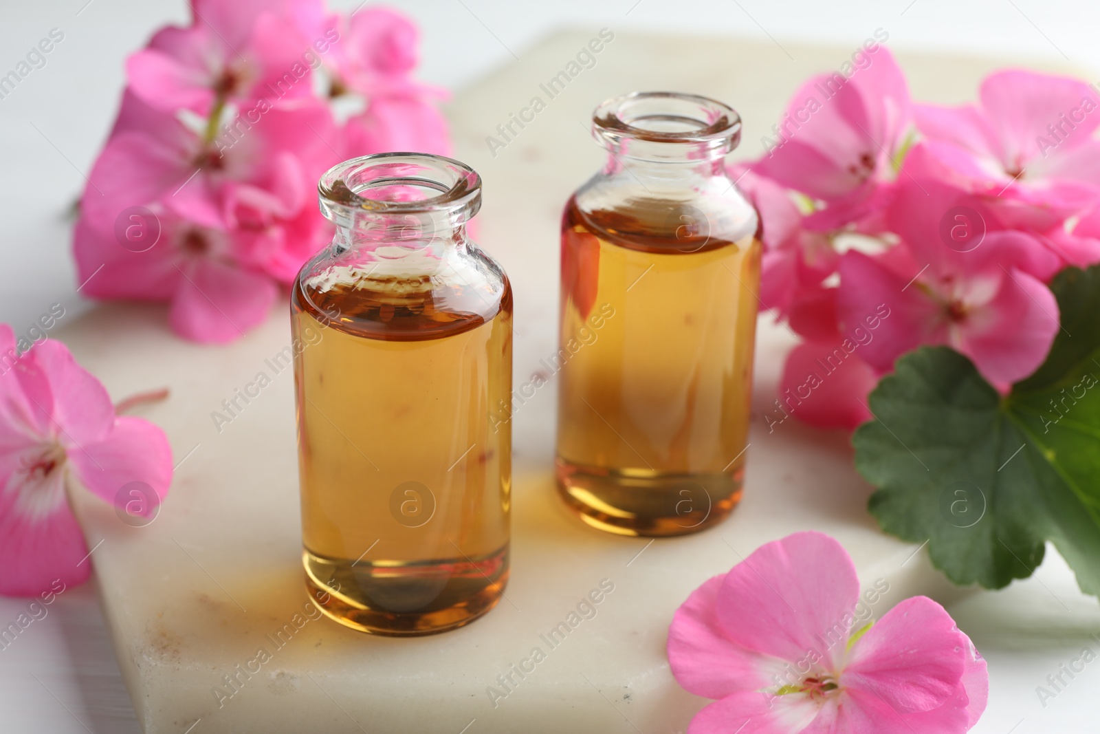 Photo of Bottles of geranium essential oil and beautiful flowers on white table, closeup