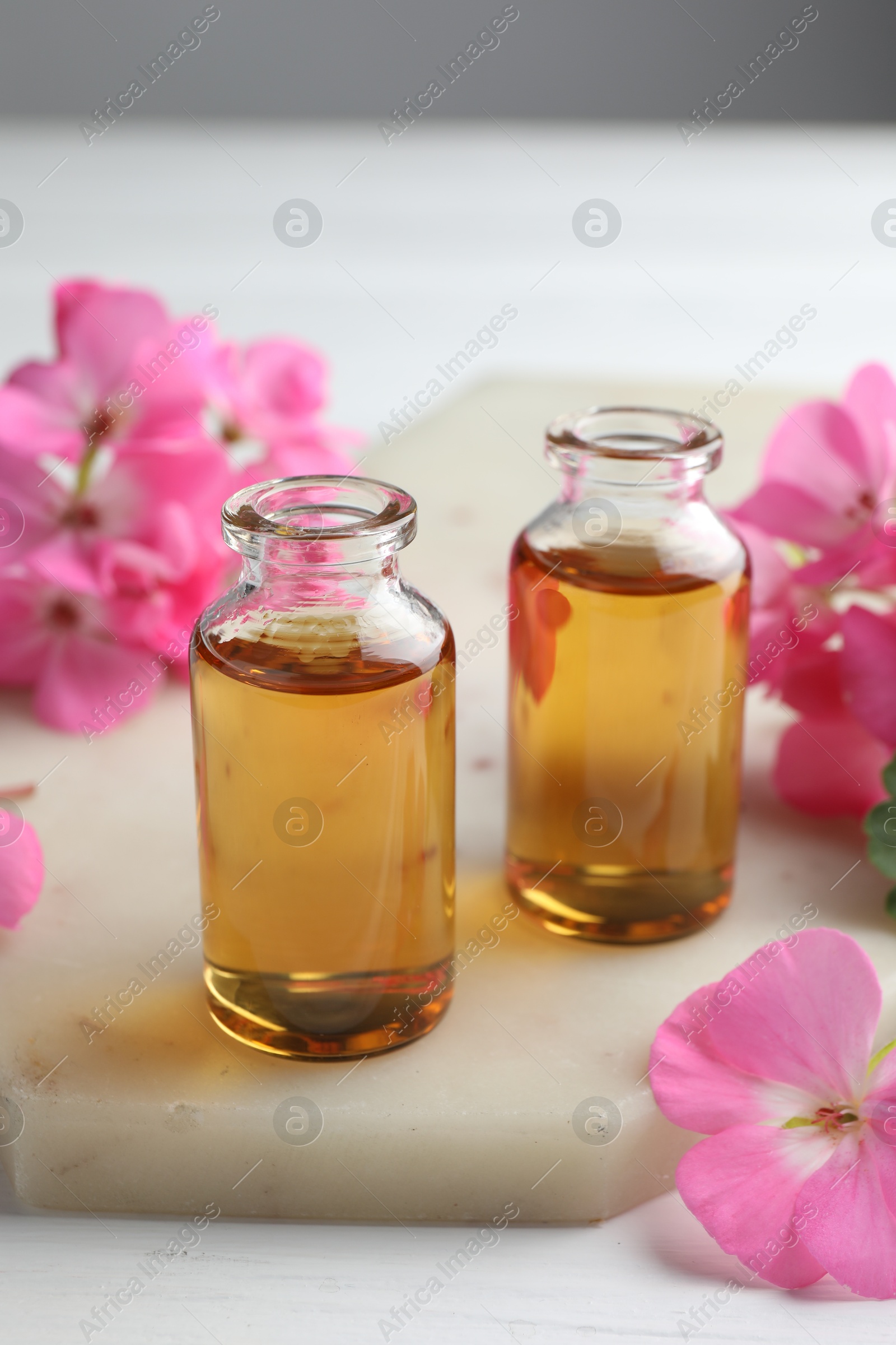 Photo of Bottles of geranium essential oil and beautiful flowers on white table, closeup