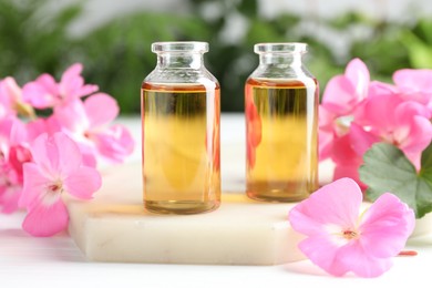 Photo of Bottles of geranium essential oil and beautiful flowers on white table, closeup