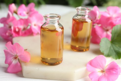 Bottles of geranium essential oil and beautiful flowers on white table, closeup