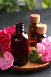 Photo of Bottles of geranium essential oil and beautiful flowers on black table, closeup