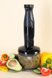 Photo of Hand blender with mixture of ingredients and fresh products on beige background