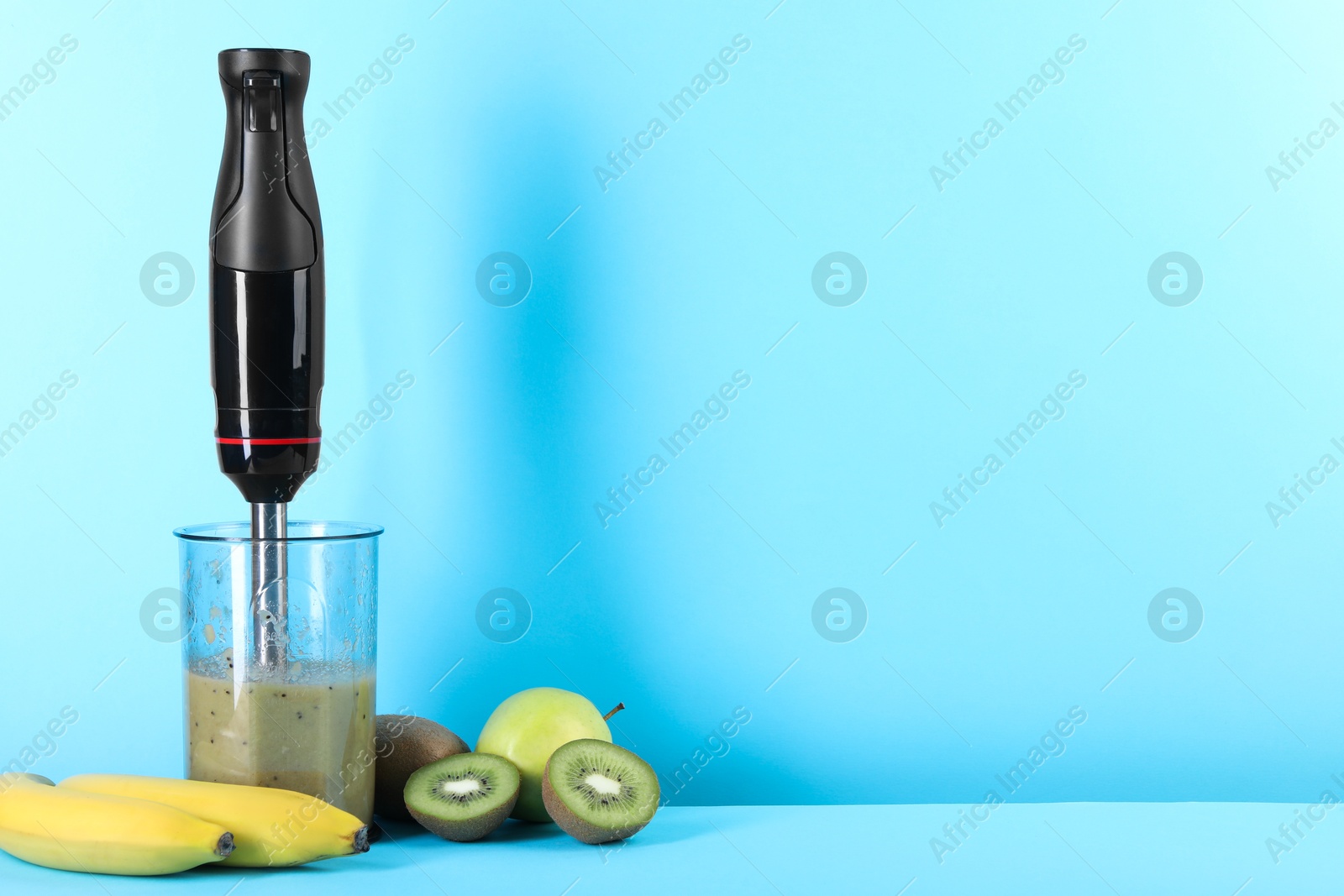 Photo of Hand blender with mixture of ingredients and fresh fruits on light blue background, space for text