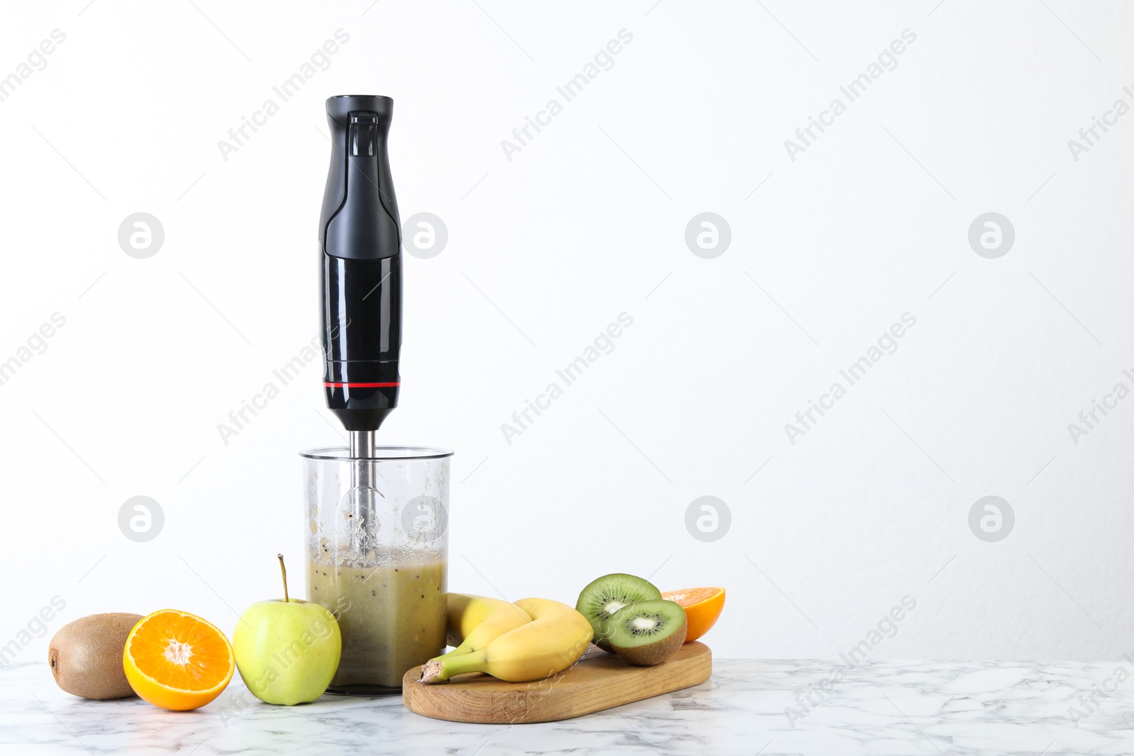 Photo of Hand blender with mixture of ingredients and fresh fruits on white marble table, space for text