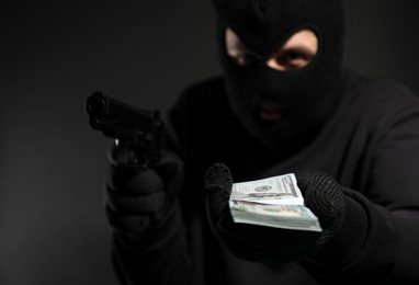 Photo of Dangerous masked criminal with gun holding money on black background, selective focus. Armed robbery