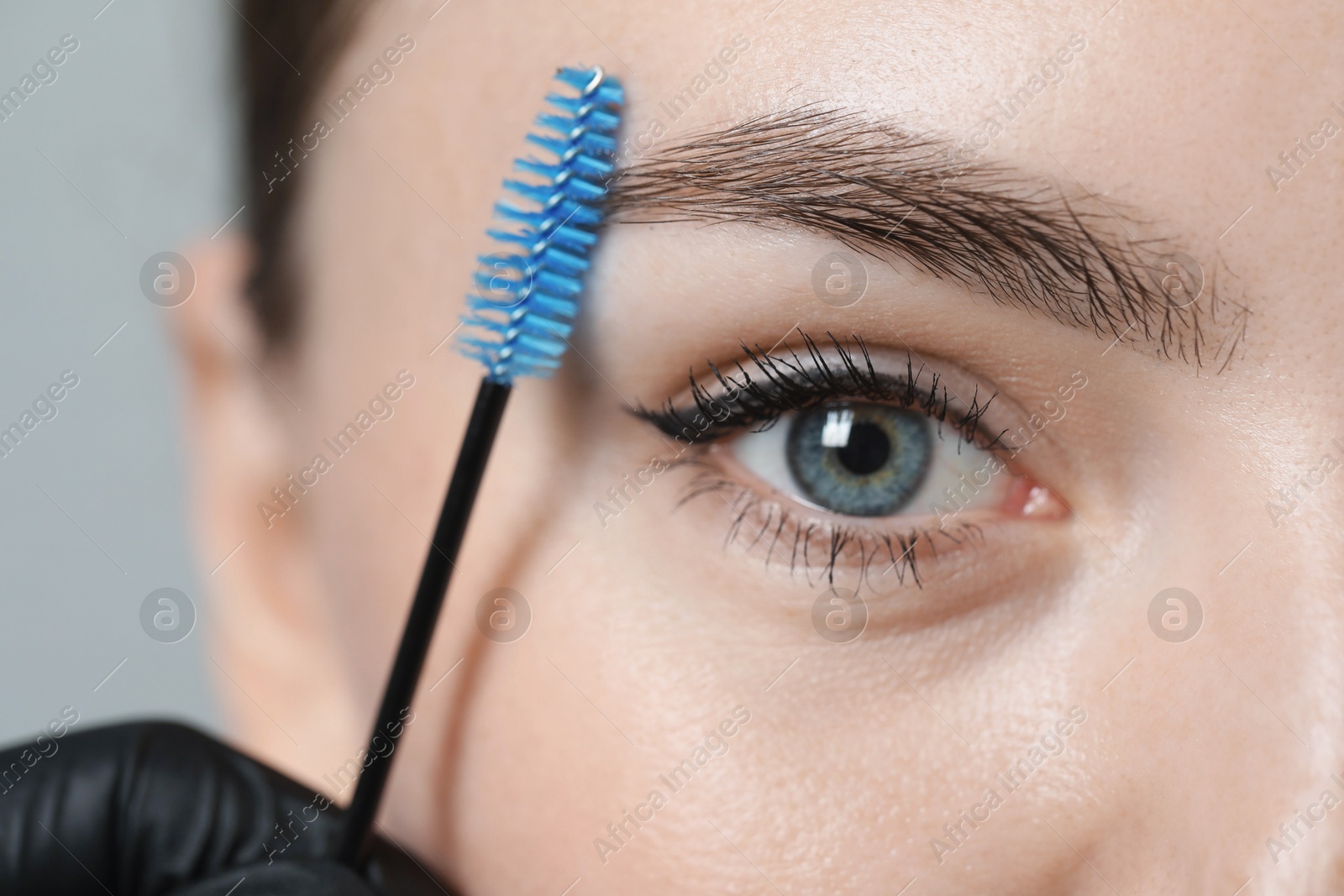 Photo of Beautician brushing young woman's eyebrow on light background, closeup