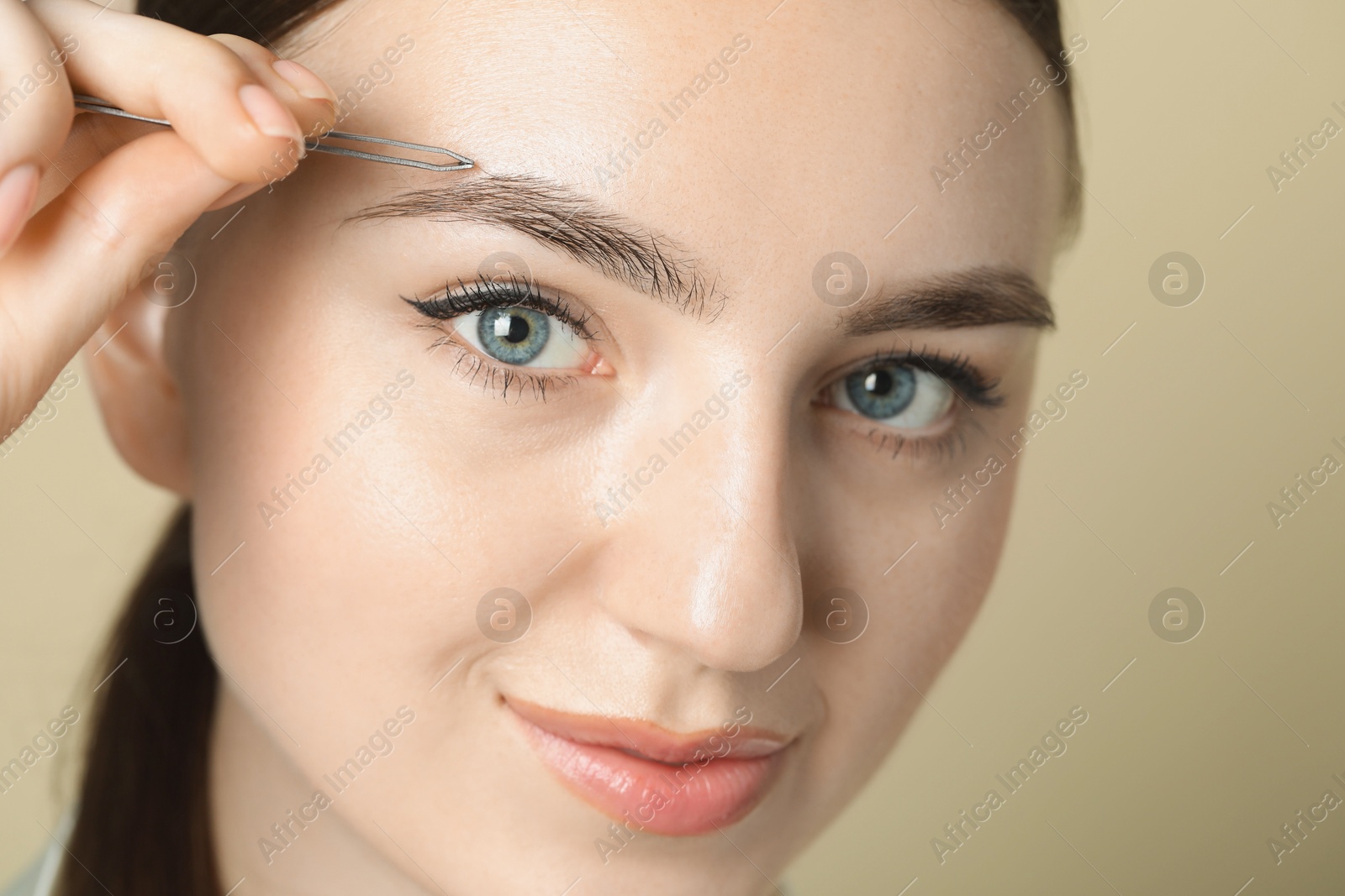 Photo of Young woman plucking eyebrow with tweezers on beige background, closeup