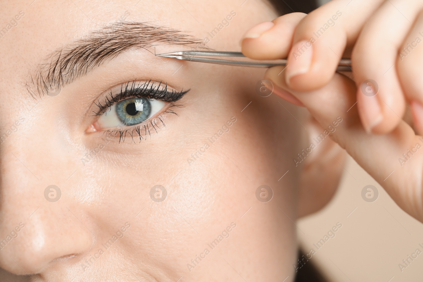 Photo of Young woman plucking eyebrow with tweezers on beige background, closeup