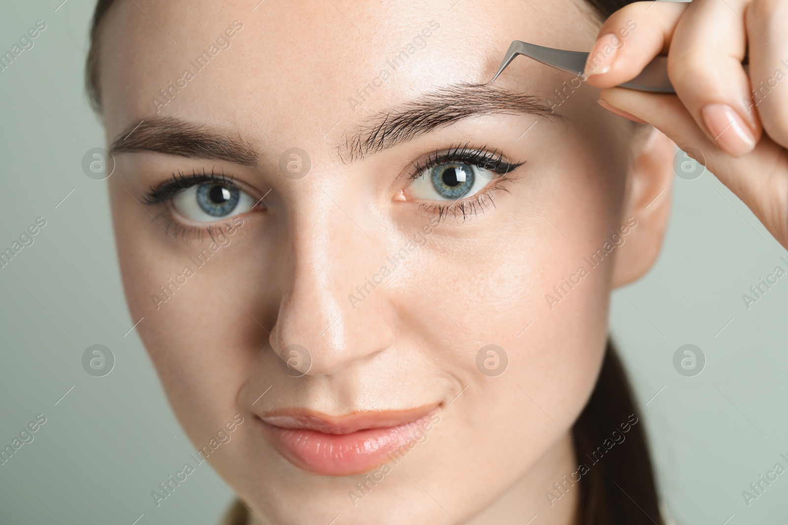 Photo of Young woman plucking eyebrow with tweezers on light background, closeup