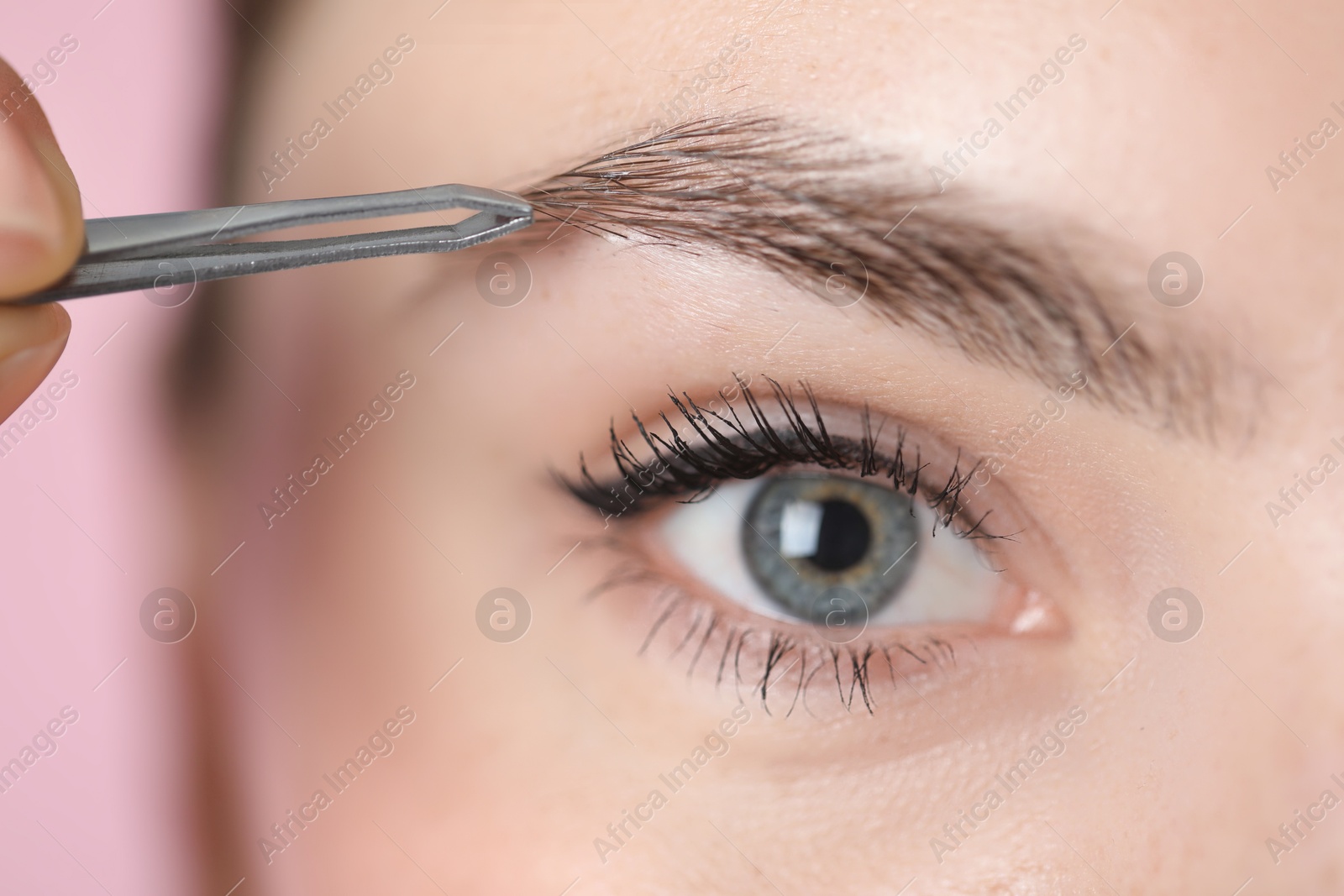 Photo of Young woman plucking eyebrow with tweezers on pink background, closeup