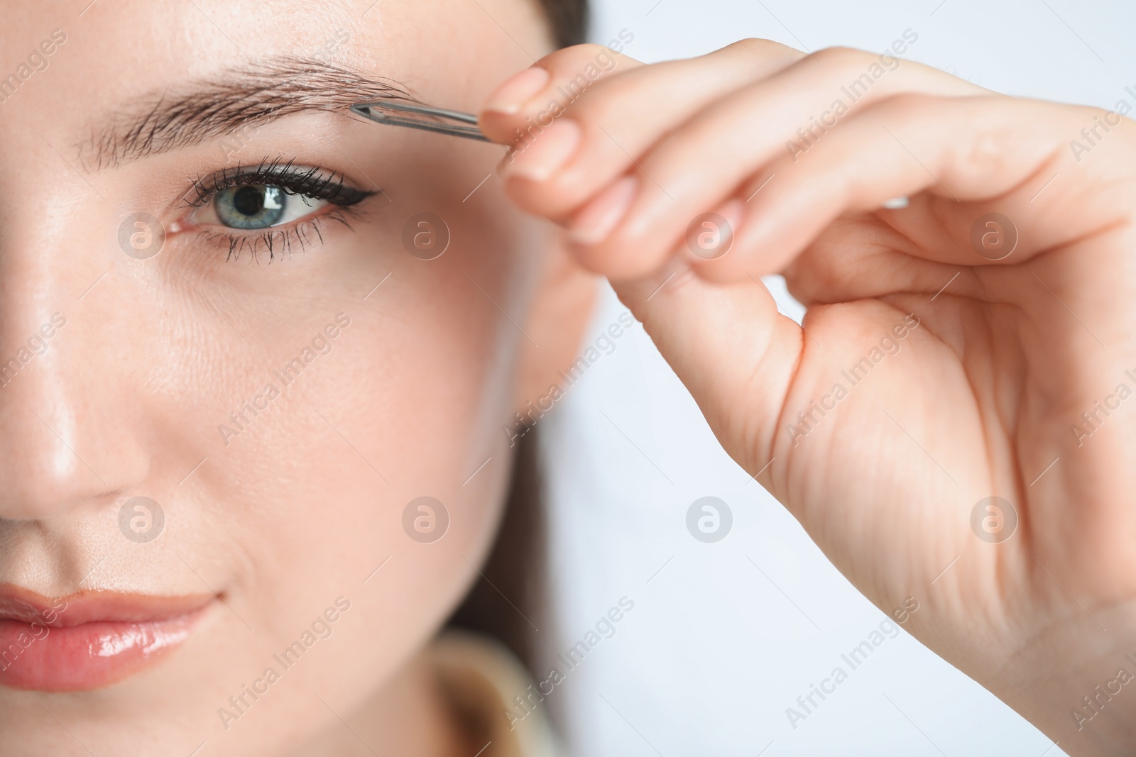 Photo of Young woman plucking eyebrow with tweezers on light background, closeup