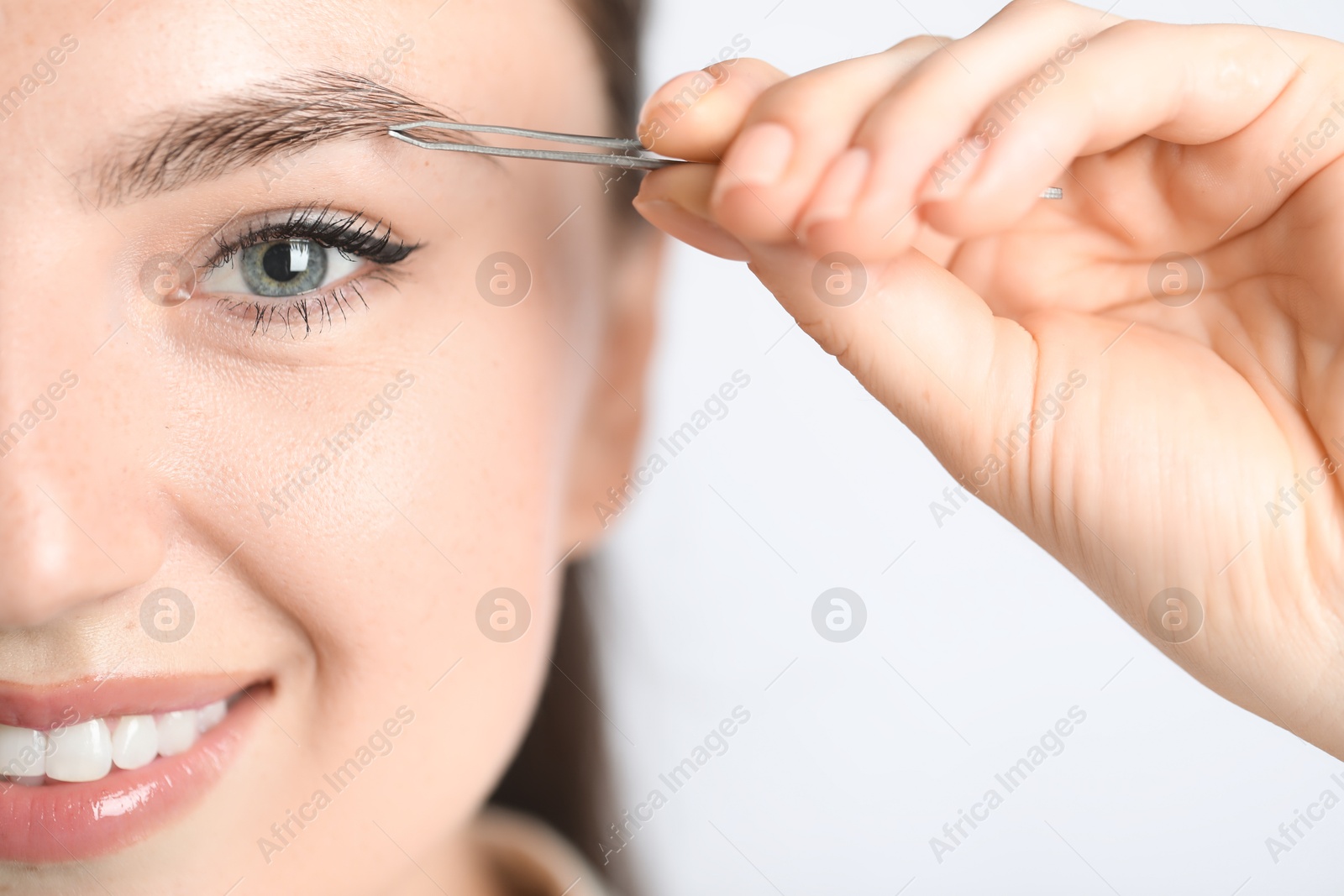 Photo of Young woman plucking eyebrow with tweezers on light background, closeup