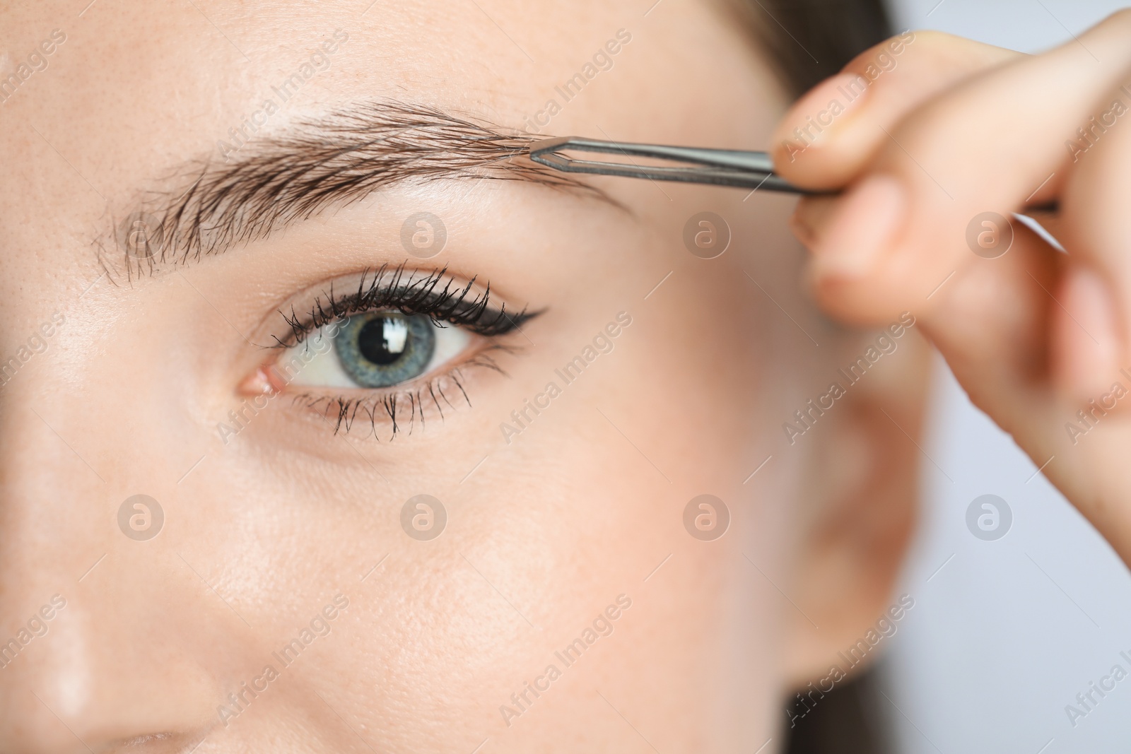 Photo of Young woman plucking eyebrow with tweezers on light background, closeup