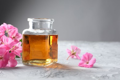 Photo of Bottle of geranium essential oil and beautiful flowers on light grey textured table, closeup. Space for text