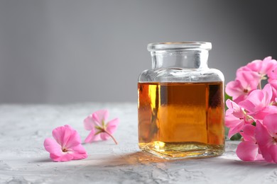 Photo of Bottle of geranium essential oil and beautiful flowers on light grey textured table, closeup. Space for text