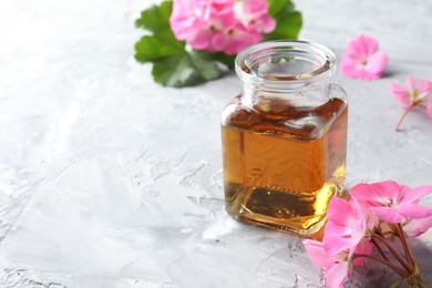 Photo of Bottle of geranium essential oil and beautiful flowers on light grey textured table, closeup