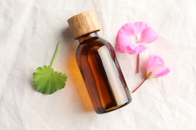 Photo of Bottle of geranium essential oil and beautiful flowers on white fabric, flat lay